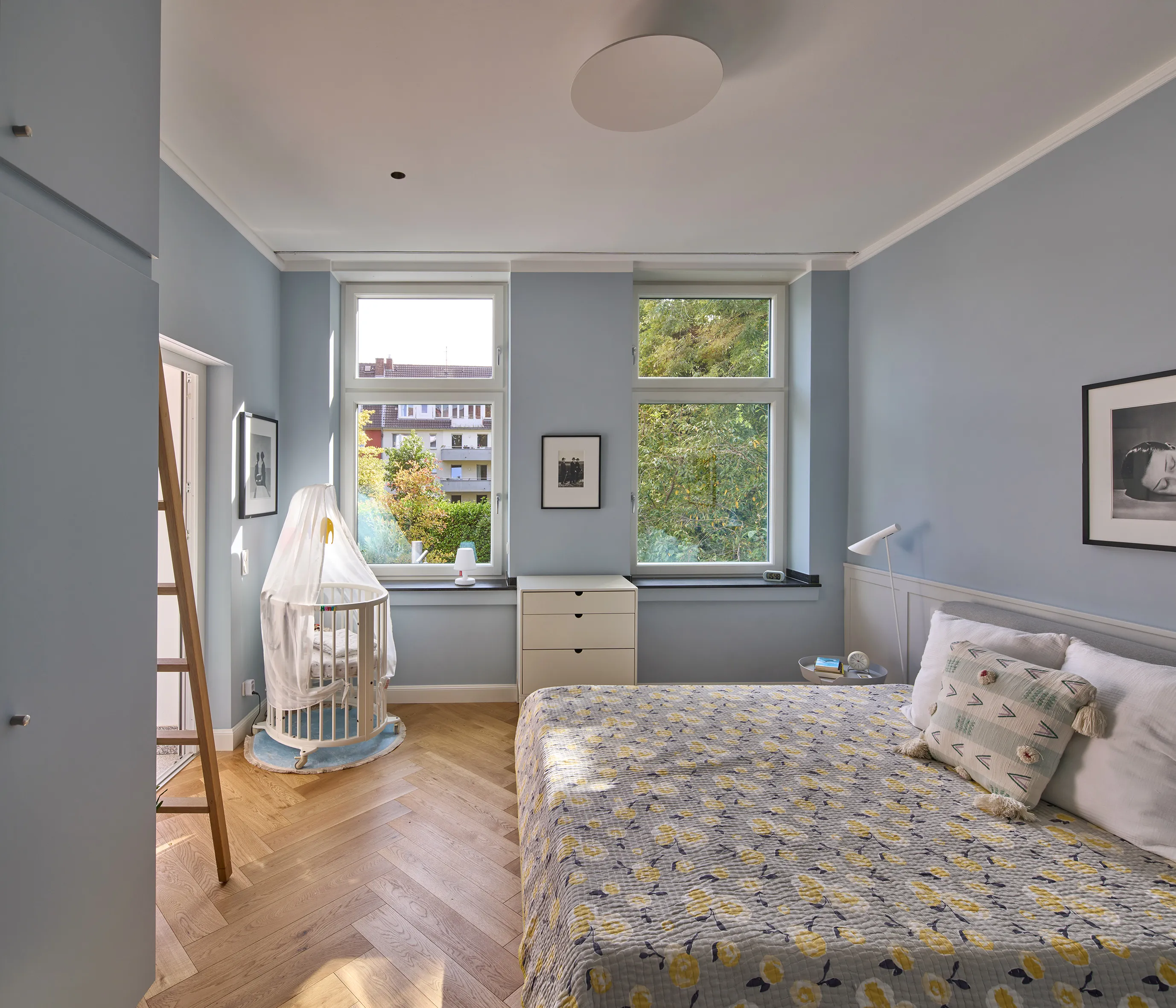 Schlafzimmer einer Maisonettewohnung mit hellblauer Wandgestaltung, großformatigen Fenstern und Eichenparkett im Fischgrätmuster.